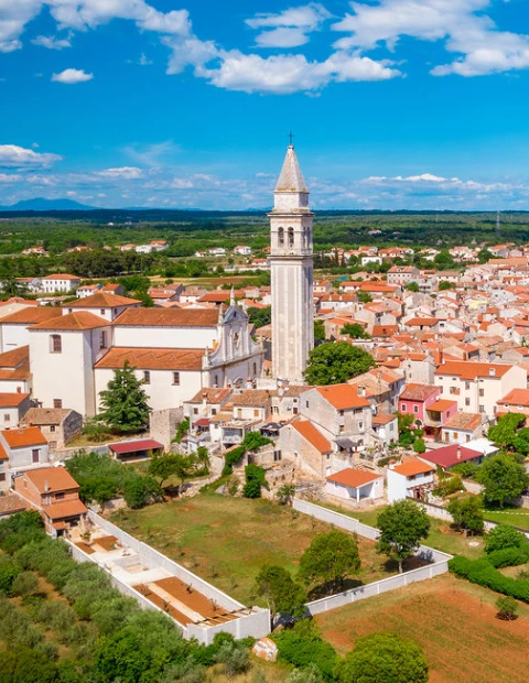 An aerial view of Galižana, a quaint village in Croatia, on a sunny day with blue skies. The image is centered on a beautiful church with a tall, slender bell tower that stands out against the surrounding landscape. The church is surrounded by tightly clustered houses with red-tiled roofs, typical of Croatian architecture. The village is nestled among lush green fields and trees, with a backdrop of gentle hills on the horizon, illustrating the idyllic rural setting of the region.