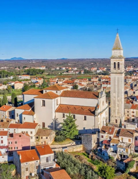 An aerial view of the city of Vodnjan, showcasing its dense historic architecture with the focal point being a large church with a tall, pointed bell tower. The church dominates the foreground, surrounded by tightly packed houses with red-tiled roofs. Beyond the urban area, the landscape opens up to show wide-open fields and scattered trees, with a mountain range faintly visible in the distance under the clear blue sky. The scene captures the blend of cultural heritage and natural beauty characteristic of this Croatian city.