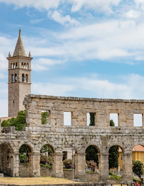 A vertical image capturing a part of the ancient Roman amphitheater in Pula, with a series of arches and stone blocks. In the background stands a medieval bell tower with a pointed spire, characteristic of the region's historical architecture. The scene is set against a partly cloudy sky, and there are hints of greenery and modern-day structures that suggest the blending of historical and contemporary elements in the city. Photo by Volker Meyer.