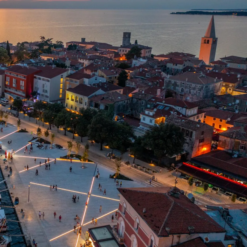 An aerial view of Poreč at twilight, capturing the warm glow of streetlights and building windows. The town square is bustling with activity, with people appearing as small figures against the expansive paved area. Two church spires rise above the historic Mediterranean architecture, silhouetted against the fading light of the sky and the Adriatic Sea in the background. The scene conveys the peaceful ambiance of a coastal town winding down for the evening.