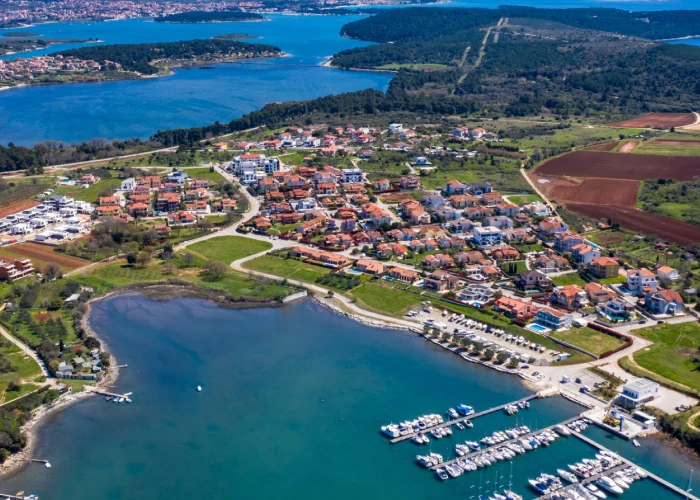 Aerial view of Banjole, a picturesque coastal village with distinctive red-roofed houses. On the left side of the image is a serene bay with clear blue waters gently embracing the shore. To the right, a marina is bustling with a variety of boats neatly docked in rows. Winding through the heart of Banjole is a road, flanked by clusters of buildings interspersed with verdant patches. Beyond the narrow channel of water, another part of the village is visible, suggesting the expanse of this charming locale. The surrounding area features a harmonious mix of lush green fields and areas prepared for agriculture, showcasing the village's blend of natural beauty and rural activity.