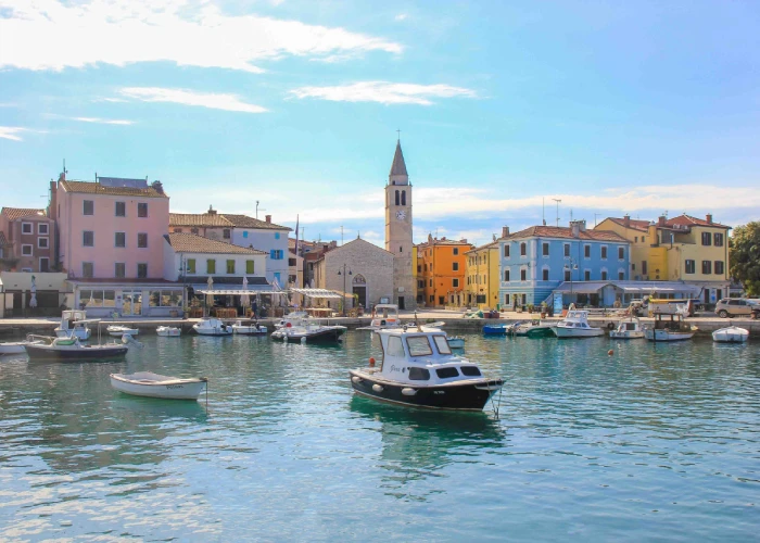 A tranquil harbor scene in Fažana, Croatia. Small boats and yachts float on the clear, calm waters in the foreground. Beyond the harbor, a row of charming multicolored houses lines the waterfront, with shades of pink, yellow, and blue reflecting the town's vibrant character. A prominent stone church with a tall, slender bell tower rises above the rooftops, providing a focal point against the soft blue sky. The overall atmosphere is peaceful and picturesque, typical of a small Croatian coastal town.