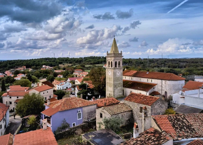 An aerial view of Ližnjan, a village in Croatia, showcasing a mix of old and new roofed houses and a prominent church with a bell tower. The church stands as a central historical landmark within the village. The landscape is marked by scattered greenery and trees, under a dramatic sky with patchy clouds that hint at the possibility of rain. The overall scene captures the rustic charm of a Croatian village steeped in history.