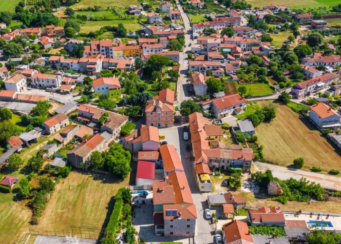 An aerial view of Marčana, a peaceful village in Croatia. The landscape is dotted with numerous red-roofed houses neatly organized along a network of streets. The architecture is a blend of traditional and modern styles, with the buildings' warm colors standing out against the surrounding greenery. Open fields and patches of land ready for cultivation border the village, emphasizing its rural charm. The tranquility of the village is palpable, with the streets showing minimal activity and the natural landscape stretching into the distance.