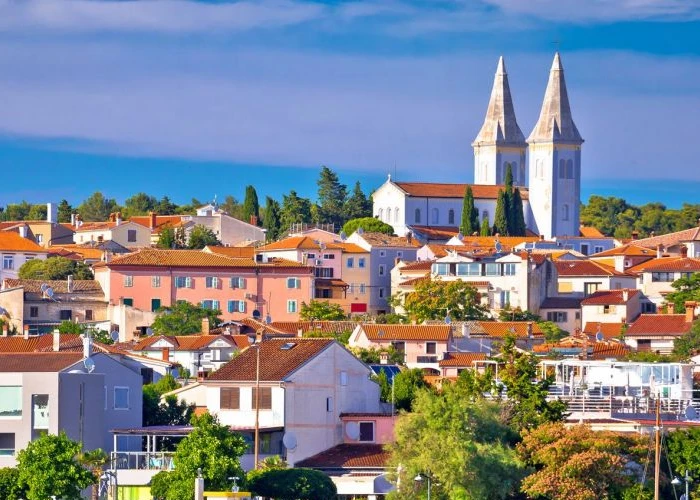 The image shows a picturesque view of a coastal town under a clear blue sky. Pastel-colored buildings with terracotta roofs are densely packed across the landscape, with lush greenery interspersed between them. Dominating the skyline are the twin white spires of a church, which stand out against the backdrop of the deep blue sea in the distance. The town exudes a Mediterranean charm, typical of coastal settlements in the region.