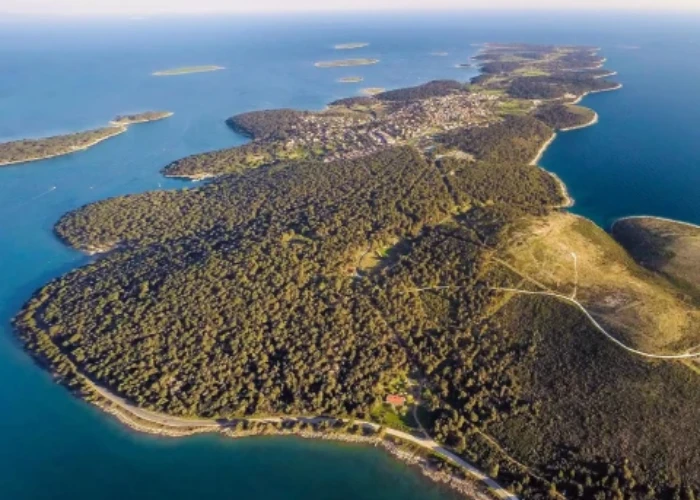 An aerial view of a verdant peninsula jutting into the calm blue waters of the Adriatic Sea. The coastline is a mix of rugged cliffs and gentle beaches, while dense forests cover the land. A clear sky above and the serenity of the isolated islands scattered in the distance convey a sense of peaceful seclusion. This landscape captures the natural beauty of the coastal region, likely around the area of Pula, Croatia.