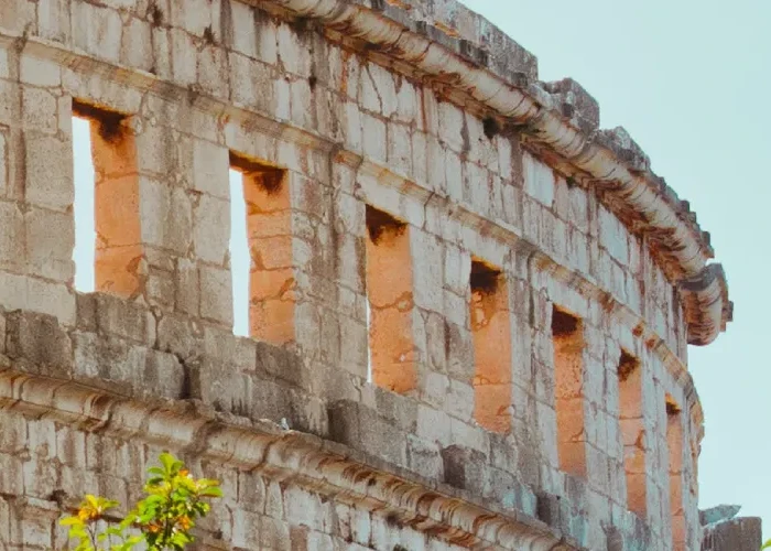 A panoramic view of the ancient Roman amphitheater in Pula, Croatia, showcasing the upper arches and remnants of the structure's facade. The weathered stone walls stand out against a clear sky, and the architecture is complemented by lush green trees peeking out from behind the ruins. Photo by Niko Pečnik.