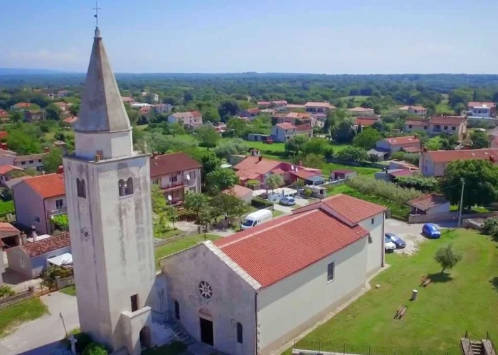 An aerial view of Šišan, a serene village in Croatia, with a church and its pointed steeple dominating the foreground. Surrounding the church are houses with red tiled roofs, nestled among lush greenery and trees, characteristic of the Croatian landscape. The image conveys a peaceful residential area with wide streets and a hint of the village's rural setting extending into the horizon under a clear sky.