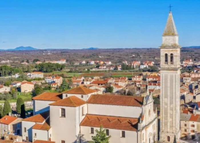 An aerial view of the city of Vodnjan, showcasing its dense historic architecture with the focal point being a large church with a tall, pointed bell tower. The church dominates the foreground, surrounded by tightly packed houses with red-tiled roofs. Beyond the urban area, the landscape opens up to show wide-open fields and scattered trees, with a mountain range faintly visible in the distance under the clear blue sky. The scene captures the blend of cultural heritage and natural beauty characteristic of this Croatian city.