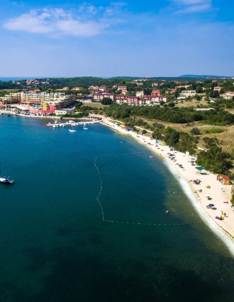 An aerial view of Banjole, a coastal area in Pula, showcasing a curved beach with people enjoying the sun and sea. The clear blue waters are calm, with several boats moored near the shore. A stretch of buoy lines marks a safe swimming area. Adjacent to the beach, a line of colorful buildings, possibly hotels and residential structures, is visible, set against a backdrop of dense greenery and trees. The area conveys a relaxed, holiday atmosphere under a bright, clear sky.