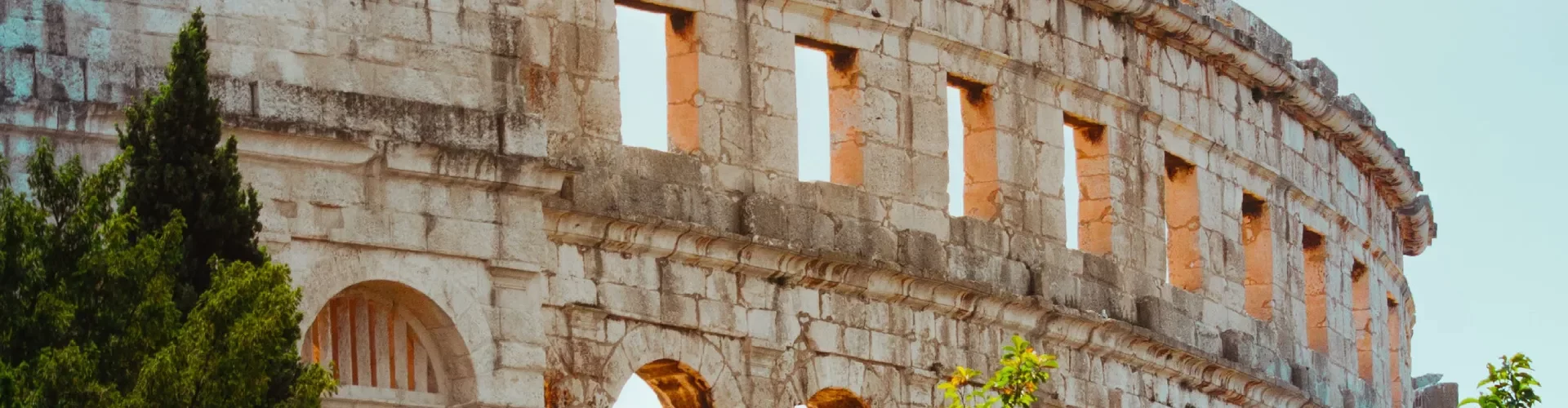 A panoramic view of the ancient Roman amphitheater in Pula, Croatia, showcasing the upper arches and remnants of the structure's facade. The weathered stone walls stand out against a clear sky, and the architecture is complemented by lush green trees peeking out from behind the ruins. Photo by Niko Pečnik.